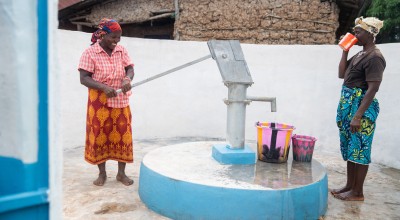 Two individuals stand at a freshly painted water pump within a walled outdoor area. One person operates the hand pump, while the other drinks from a cup. Several colourful buckets are placed on the ground near the pump, and a building made of natural materials is visible in the background.