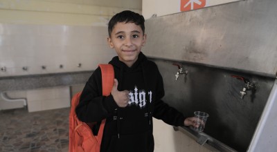 A child wearing a dark hoodie and carrying an orange backpack stands at a metal water station, holding a small plastic cup under one of the taps. The background shows a tiled floor and additional sinks along the wall.