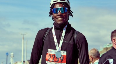 A smiling participant wearing a helmet, sunglasses, and a medal stands outdoors after completing the race, with a numbered bib pinned to their chest.