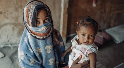 An adult seated indoors holding a young child on their lap. The adult wears a patterned blue garment and a face covering that leaves only the eyes visible. The child wears a light-colored outfit and has short hair tied in small sections. The setting appears to be a simple interior space with textured walls and bedding visible in the background under soft natural light.