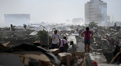 People walk through the aftermath of widespread destruction in a heavily damaged urban area, where collapsed buildings, debris, and smoke stretch across the landscape. A few damaged high-rise buildings remain standing in the background as residents and responders move carefully through the ruins.