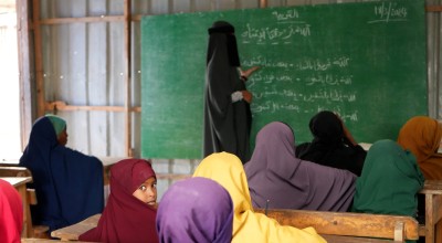 Teacher writing on chalkboard while children sit at desks in a classroom.