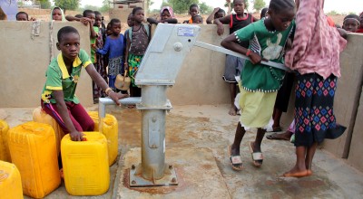 Children gather around a communal hand-operated water pump in an outdoor rural setting. In the foreground, one child in a green shirt crouches beside several large yellow plastic jerrycans, holding the spout as clear water flows into one container. Another child to the right is pushing the metal pump handle up and down to draw water. Behind a low concrete wall, a group of children stand closely together watching, some leaning over the wall. The ground around the pump is wet with small puddles, and the pump 