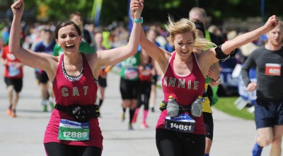 Two women runners in matching bright pink vests celebrate with arms raised and joyful expressions as they approach the finish line of a marathon. The runner on the left wears bib number 12889 with "CAT" on her vest, while the runner on the right wears bib 14681 with "ANNIE" on hers. Other participants and spectators are visible in the background under a sunny sky.