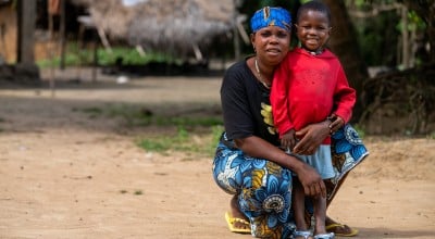 A woman wearing a blue patterned headscarf and patterned wrap skirt crouches beside a smiling young child in a red hoodie on a sandy path. They face the camera together in front of a thatched-roof home, with trees and simple village buildings softly blurred in the background.