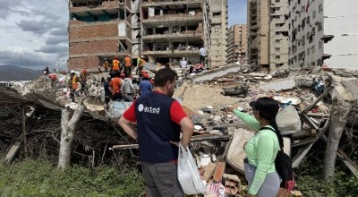 Aid worker and resident stand beside rubble as rescue teams search debris in front of a heavily damaged apartment building.