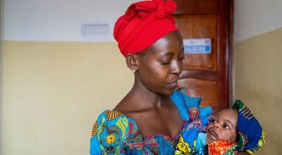 A woman wearing a bright red headscarf and colourful patterned dress cradles her baby in her arms while looking down with care. Wrapped in a vibrant blanket, the baby looks back up at her inside a healthcare facility.