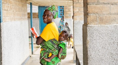 A smiling mother wearing a bright yellow top and green patterned wrap carries her young child on her back while holding a sachet of nutrition supplement. She looks back toward the camera as they walk through a health centre corridor, with other families visible in the background.