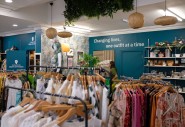Charity shop interior with clothing racks, a donation drop-off point, homeware shelves, hanging plants, and a wall sign reading, “Changing lives, one outfit at a time.”