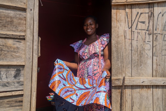 A smiling woman in a pink patterned dress stands in a wooden doorway while holding a piece of fabric.