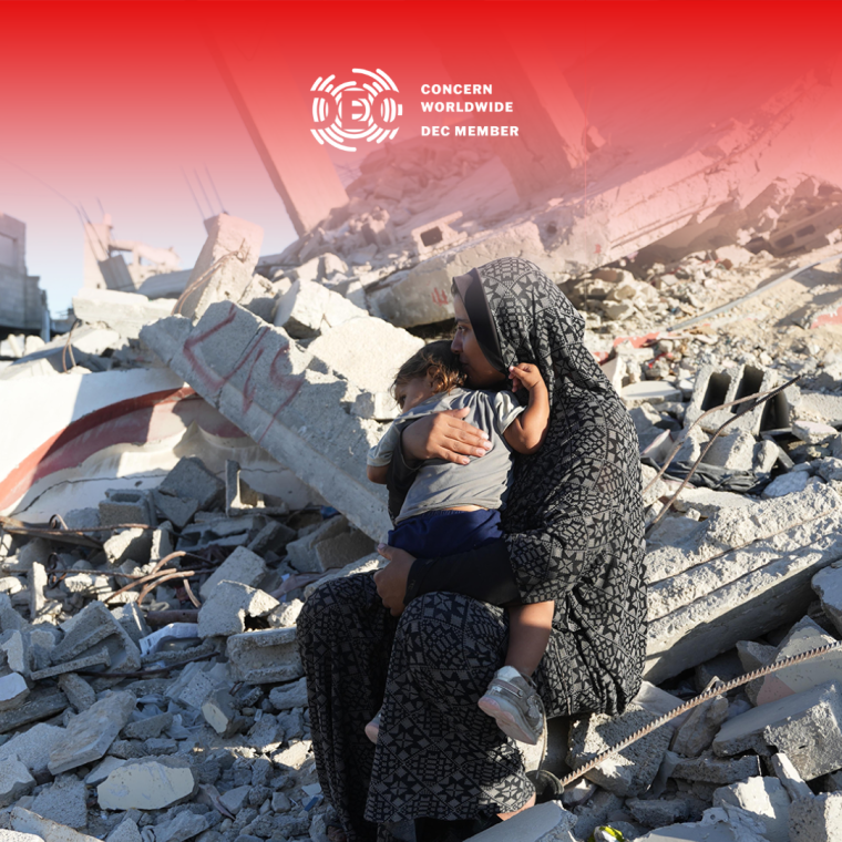 A woman in a patterned robe sits among the rubble of a destroyed building, holding a young child in her arms. The background shows debris and collapsed concrete.