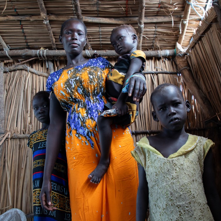 A woman stands inside a shelter made of sticks, holding a small child on her arm. Two other children stand close beside her, looking directly at the camera. The woman wears a brightly patterned orange and blue dress, and the children are dressed in colorful clothes.