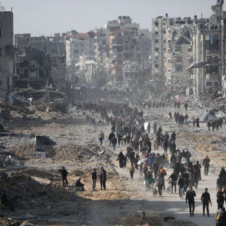 Crowds of people walk through a wide street filled with rubble and debris, flanked by heavily damaged and destroyed buildings in a war-torn city.