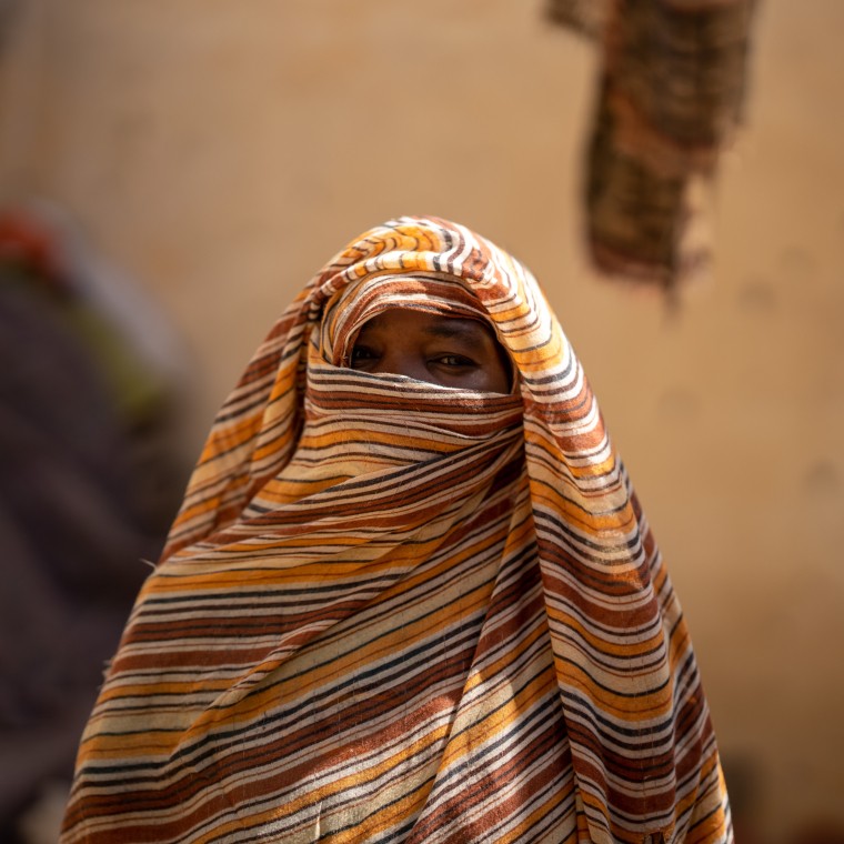 A woman stands outdoors with her face partially covered by a striped headscarf, leaving only her eyes visible. She looks calmly toward the camera, with a softly blurred background of earth-toned walls and figures behind her.