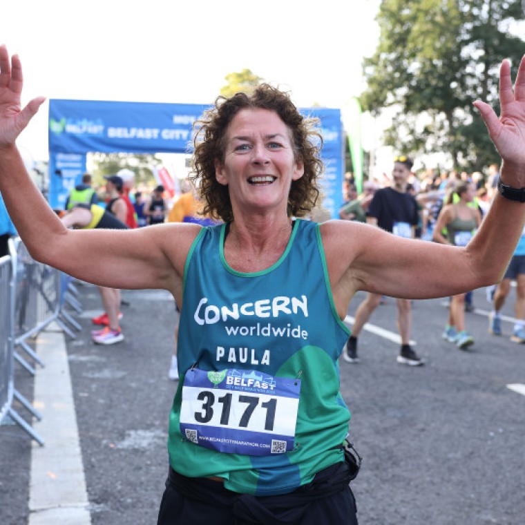 A woman wearing a Concern Worldwide running top holds her hands up and smiles as she runs a race