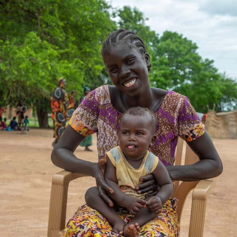 A mother sits on a charity with her happy baby on her lap. Behind her are people in colourful clothing and large green trees. 