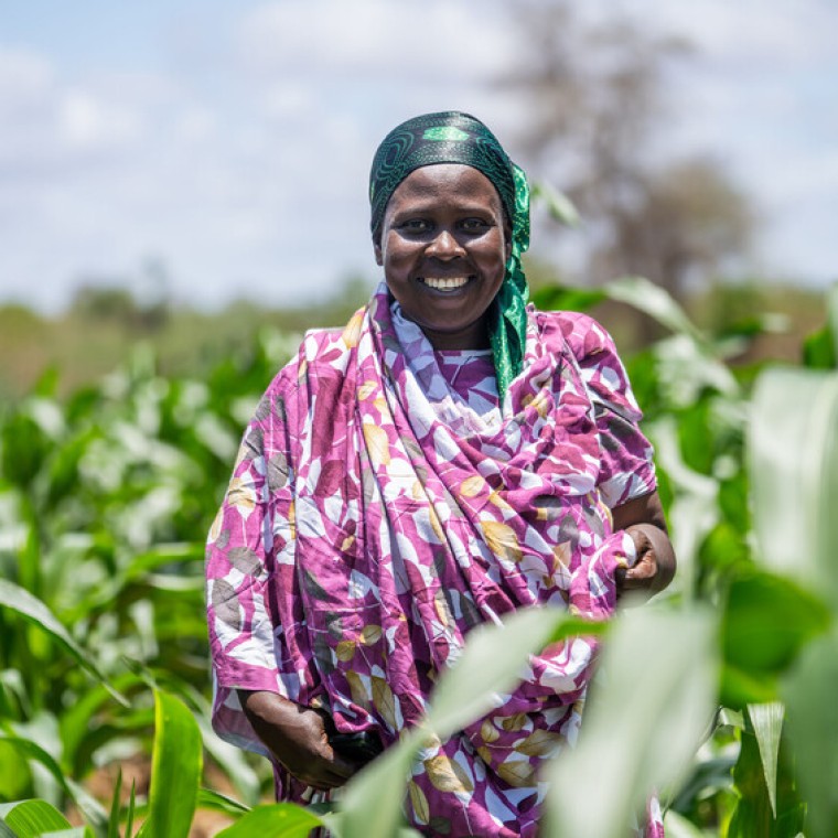 A smiling woman wearing purple stands in a field surrounded by plants.