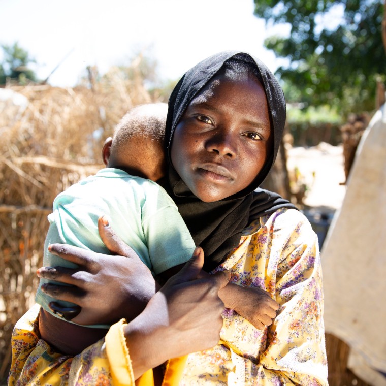 A young woman in a headscarf looks into the camera while holding her young baby. There is a rural outside background behind her.