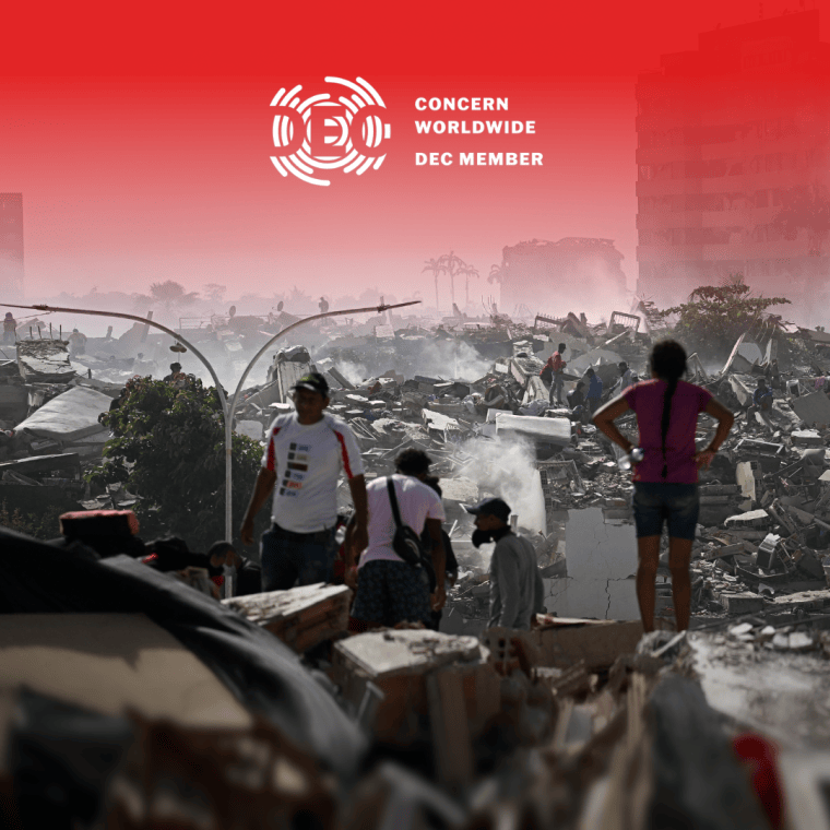 People walk through the aftermath of widespread destruction in a heavily damaged urban area, where collapsed buildings, debris, and smoke stretch across the landscape. A few damaged high-rise buildings remain standing in the background as residents and responders move carefully through the ruins.