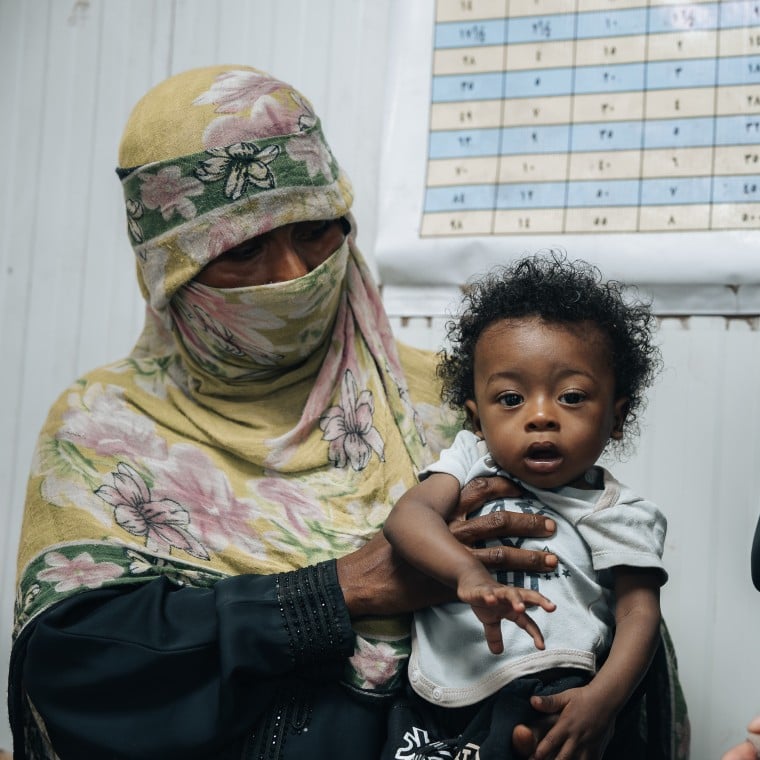 A woman wearing a patterned face covering holds a young child while standing inside a healthcare or community support centre. Another woman wearing a black face veil stands nearby, and a chart is visible on the wall in the background. The child looks toward the camera as the adults participate in what appears to be a health, nutrition, or support service visit. The image highlights maternal and child healthcare support in a community setting.