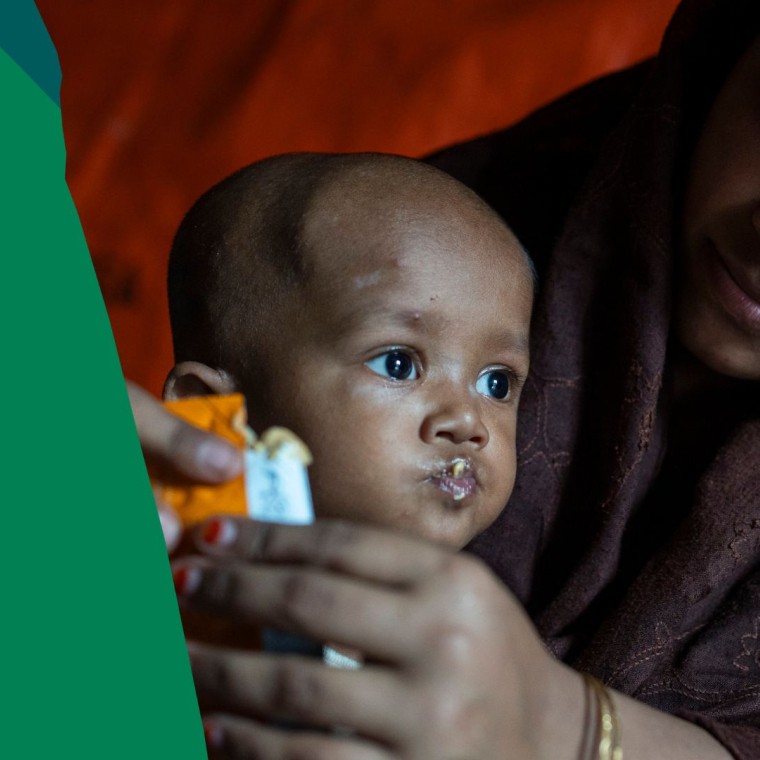 Close-up of a mother wearing a dark headscarf holding and feeding a young child inside a dimly lit shelter or tent. The child is eating from a food sachet held in the mother’s hands.  