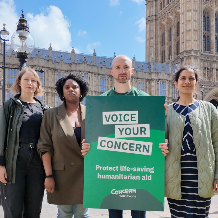 Four campaign supporters stand outside the UK Parliament holding a large green sign that reads “VOICE YOUR CONCERN – Protect life-saving humanitarian aid.” The group faces the camera in front of the Palace of Westminster on a sunny day, highlighting a public advocacy campaign calling for the protection of humanitarian aid funding.