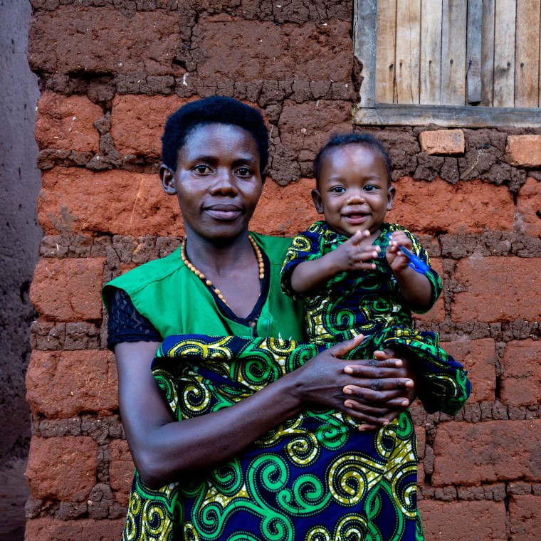A woman holds a smiling young child outside a home built from mud bricks. Both wear matching green patterned clothing and look toward the camera, while the child smiles and holds a small blue object. A wooden shuttered window is visible behind them.